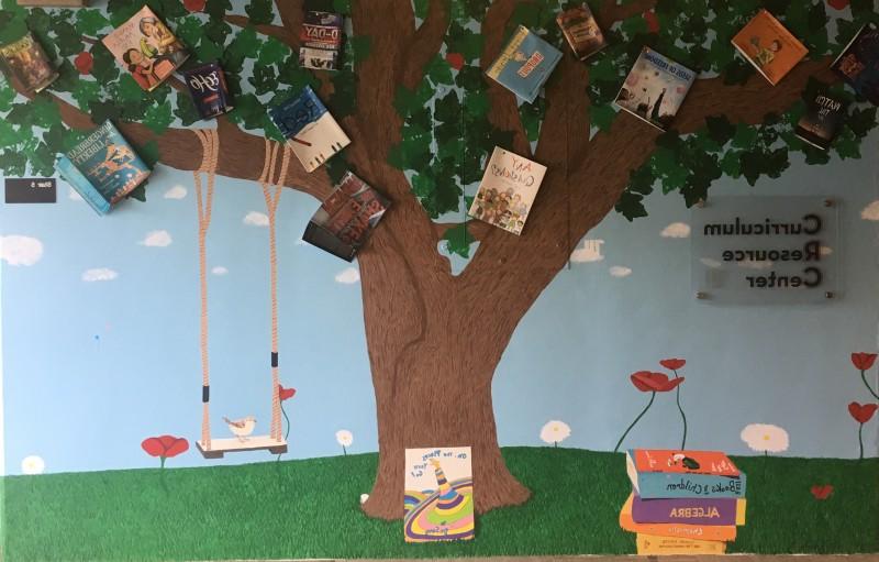 CRC Mural: depicts a tree in a field. There are book covers scattered among the leaves. There is a swing hanging from a branch, and a bird perched on the swing. A stack of books sits next to the tree, and a frog sits atop the books.